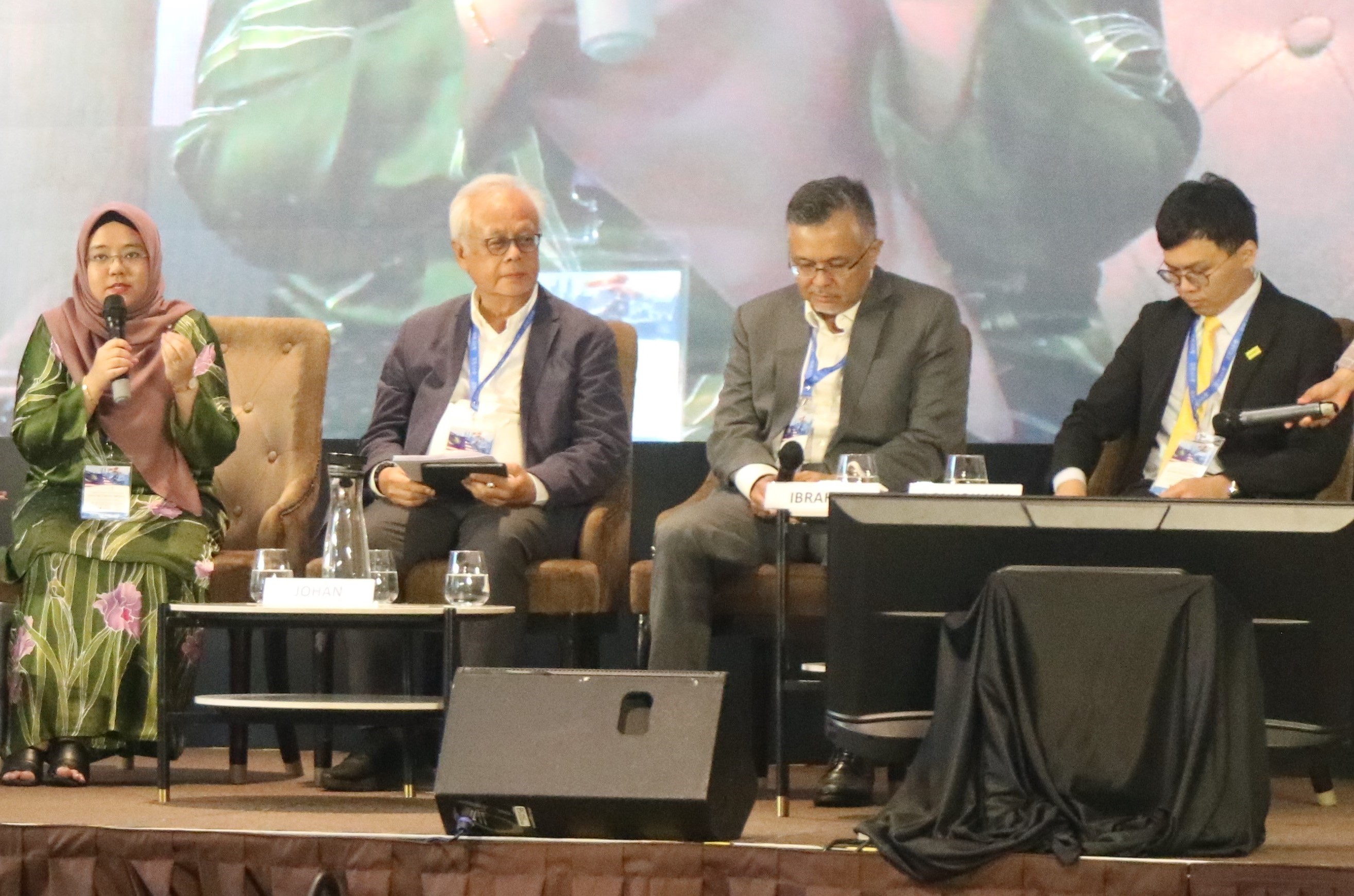 A photo of one of the sessions in Malaysia Outlook Conference 2024. Four people are sitting on individual chairs in front of a big screen on the stage. They are: Aira Azhari (Moderator), Ibrahim Suffian (Panelist), Ooi Kok Hin (Panelist), and Datuk Dr. Johan Arriffin Samad (Panelist).