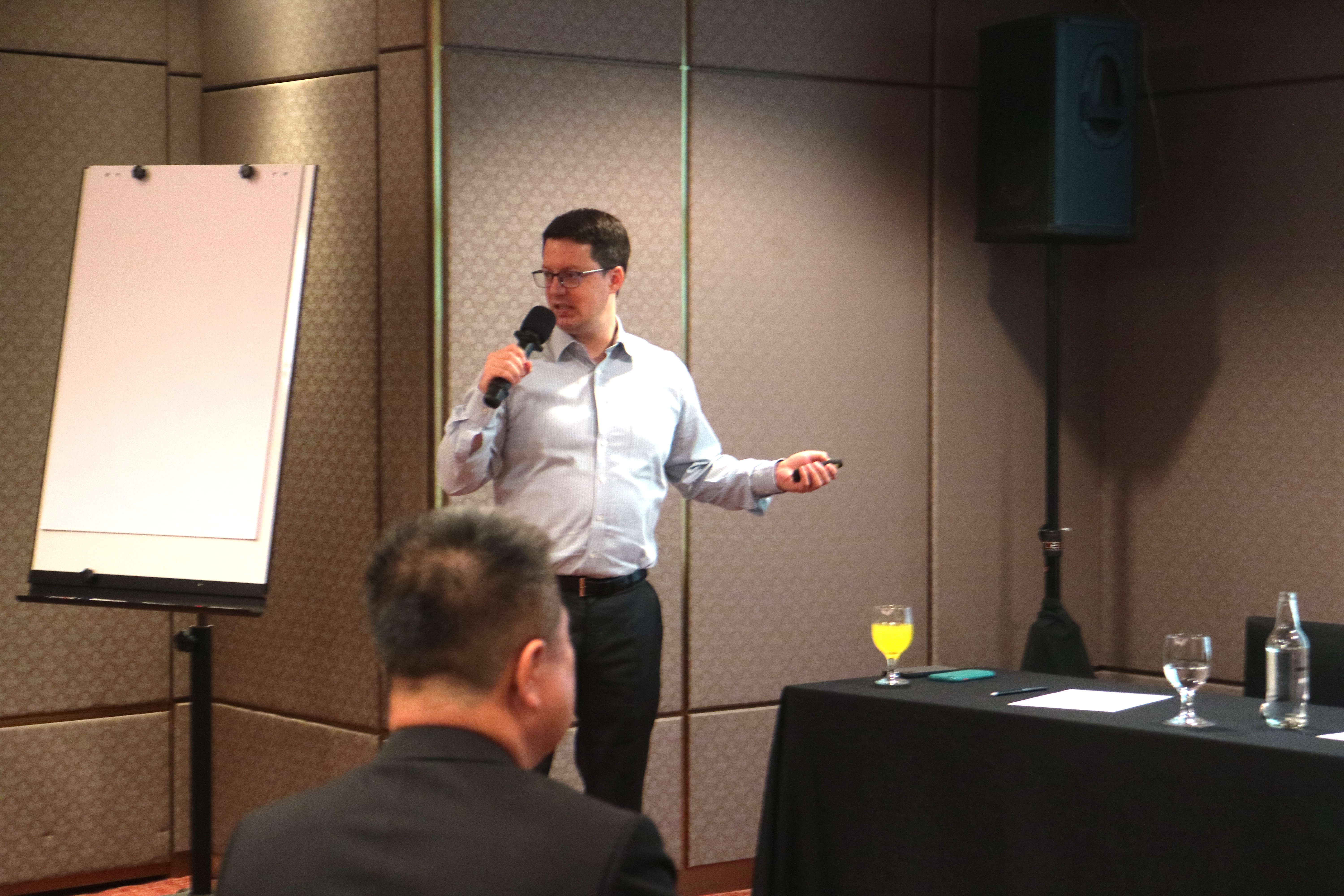 Dr. Stefan Diederich, holding a mic, in front of the room. On his left is a blank flipboard. On his right is a black table with glasses on top.