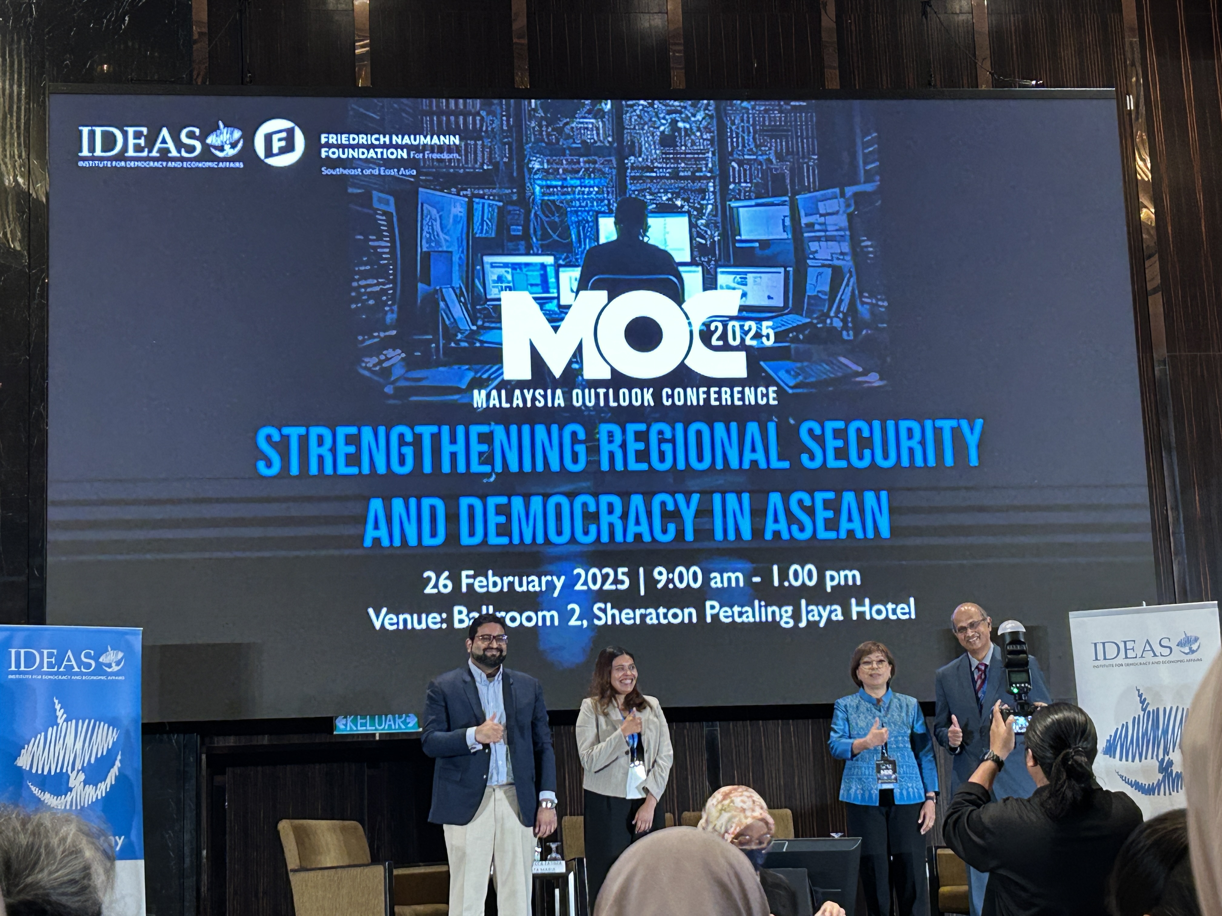 A picture from the stage of Malaysia Outlook Conference 2025. Four people (3 panelists and 1 moderator) are posing for a picture in front of the event banner.