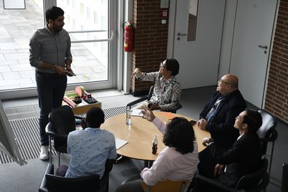 Hafizh Nabiyyin (white shirt with glasses) sitting on a round table with four other people. Hafizh is seen discussing something with someone standing next to him.