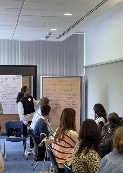 Kasturi is presenting something on the topic of Facilitation and Program Design.  She is standing in front of the room, facing the other participants. On the paper board she is showing is written words and sentences such as, "conservative"; "a retired professor respected in his field; "progressive"; "a kid of digital revolution", "human rights activists, famous blogger, and influencer".
