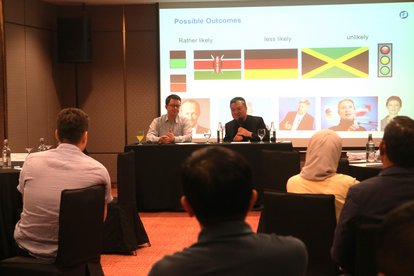 Dr. Stefan Diederich (left) sitting with Professor Wong Chin Huat (right) at a black table in front of the room, facing the participants. Behind them is a presentation slide from Dr. Diederich's PPT, showing Germany's party coalitions through colours.