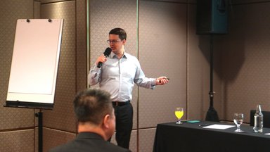 Dr. Stefan Diederich, holding a mic, in front of the room. On his left is a blank flipboard. On his right is a black table with glasses on top.